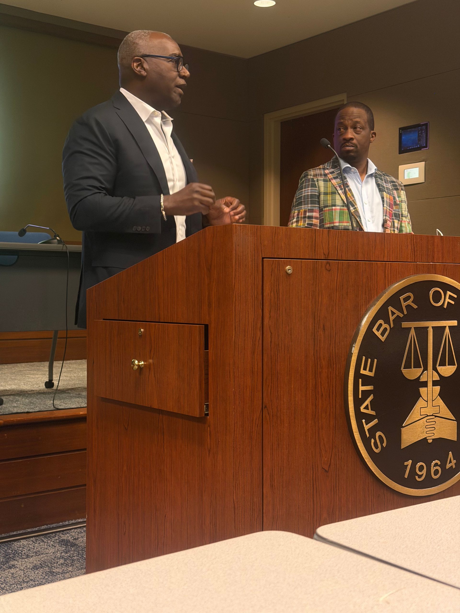 Two men stand at a podium in a government chamber, one speaking and one listening.