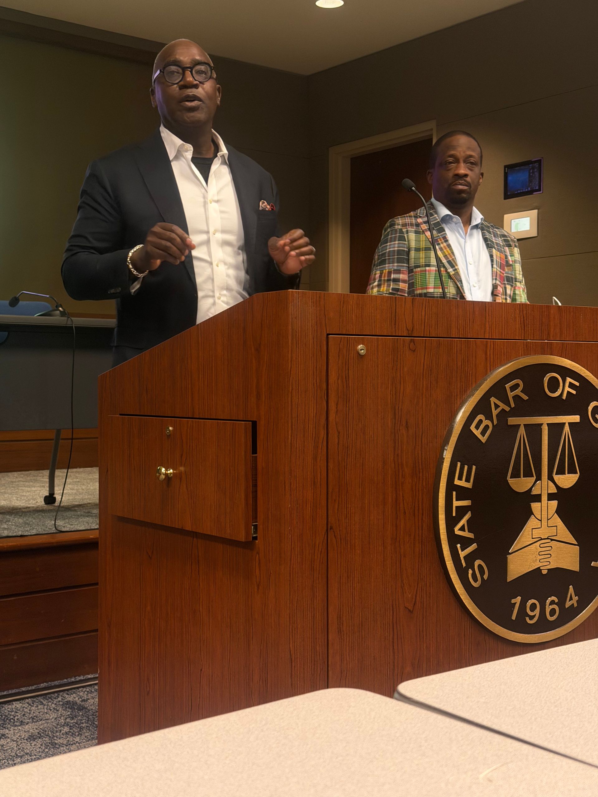 Two men standing behind a podium in a courtroom, one speaking and one looking on.