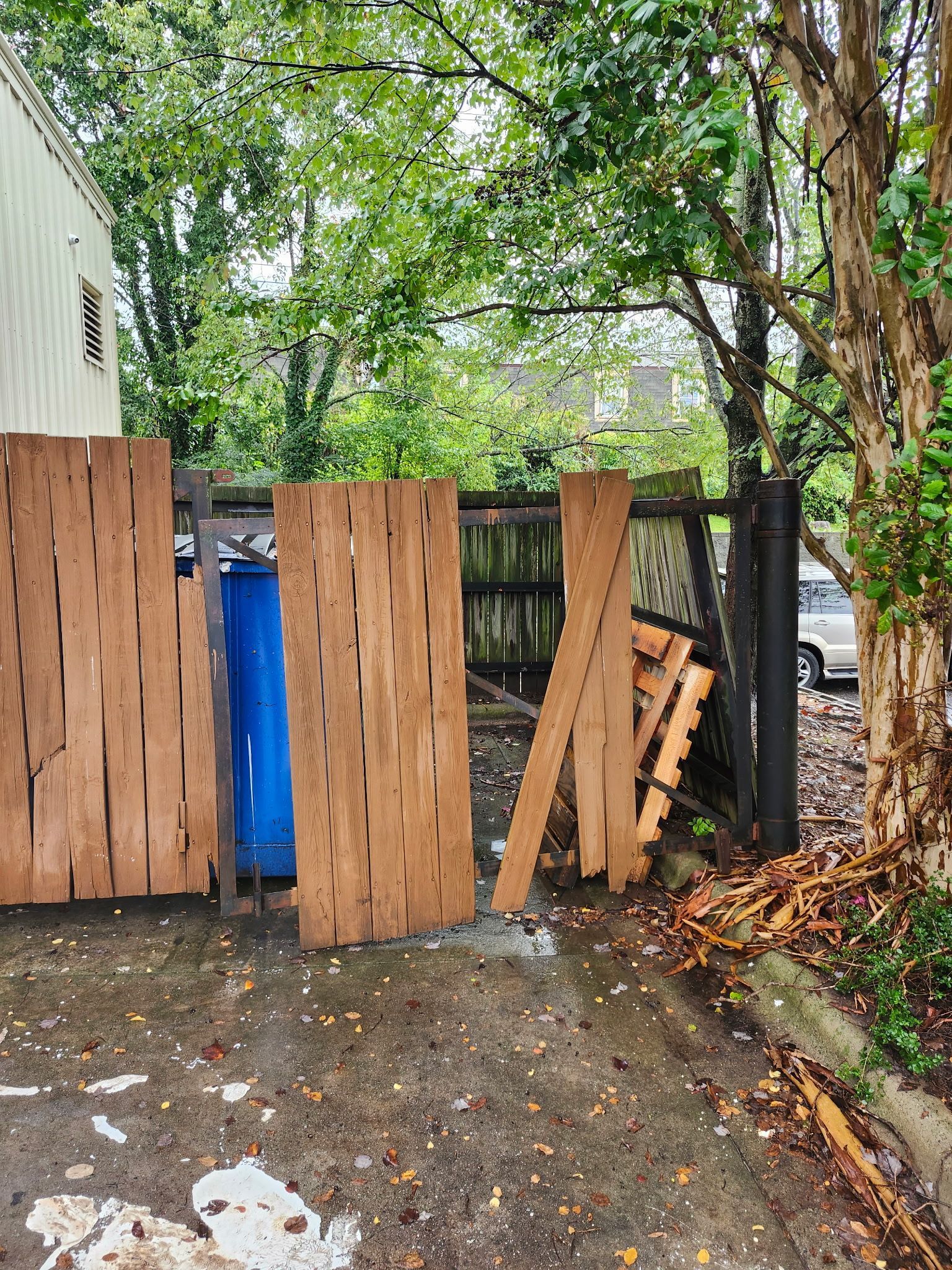 A partially dismantled wooden fence with scattered boards and a blue container in a backyard setting.