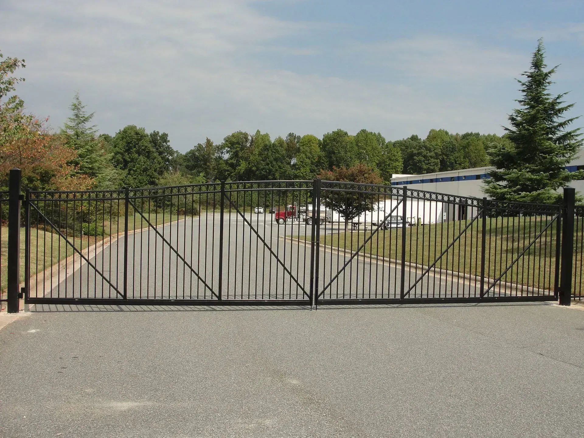 Black metal gate, closed, on gravel driveway leading toward a building and trees under a blue sky.