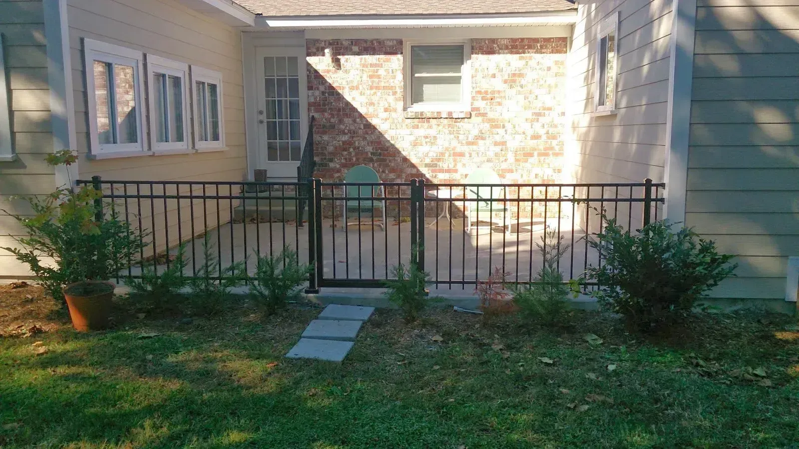 Black metal fence surrounds a small patio area with a gate. Two bushes flank the fence; grass and a pathway in front.