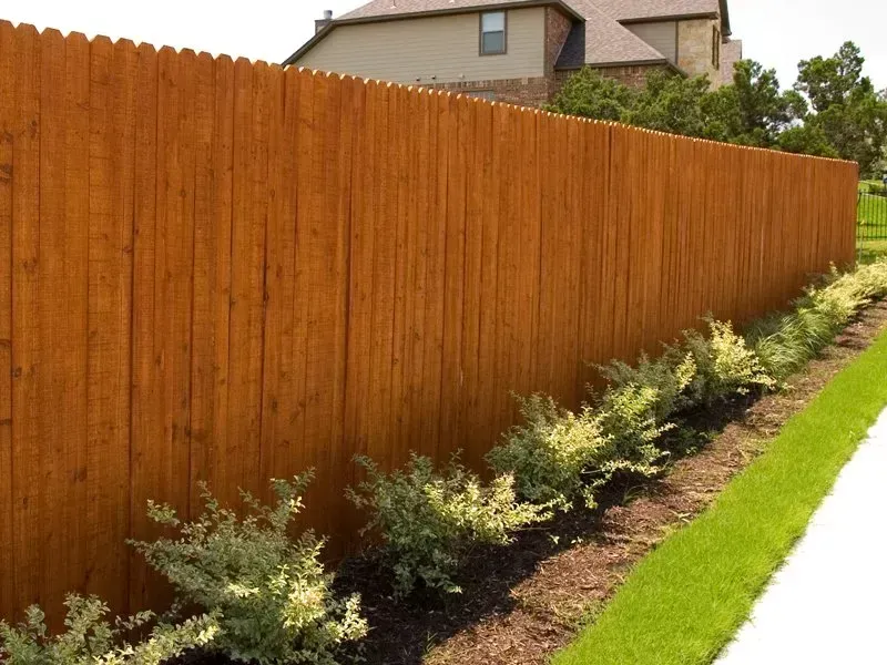 Wooden fence, stained brown, along a green lawn with small bushes and a sidewalk.
