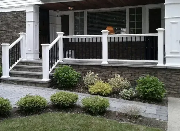 Stone-faced house porch with white railings, steps, and black spindles. Green bushes line the walkway.