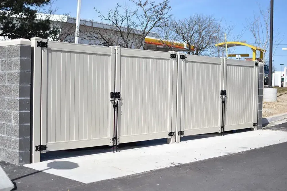 Tan vinyl dumpster enclosure with black hardware, set between gray block pillars, concrete pad.