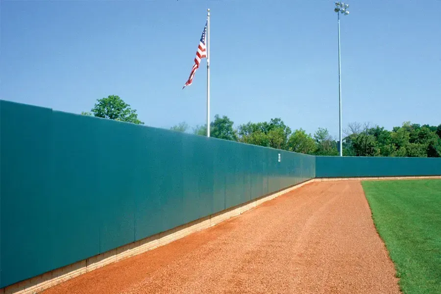Green outfield wall in baseball field with American flag and blue sky.