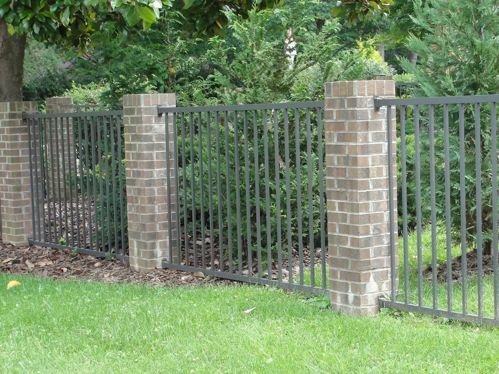 Brick pillars support a dark metal fence along a green lawn with shrubs in the background.