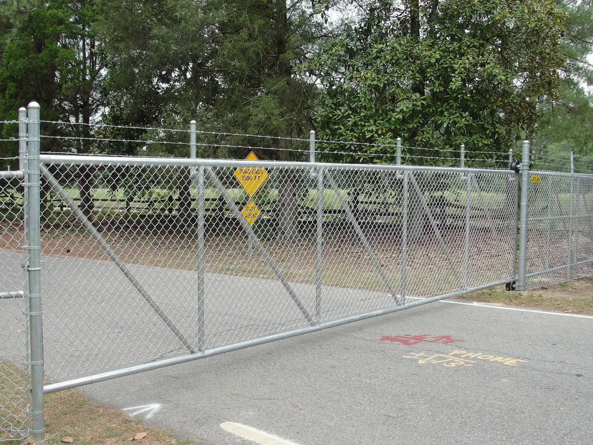 Chain-link fence with barbed wire on top, blocking a road. Trees are in the background.