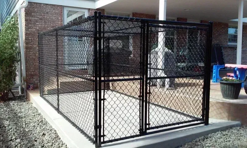 Black chain-link dog kennel on concrete patio next to a brick building.