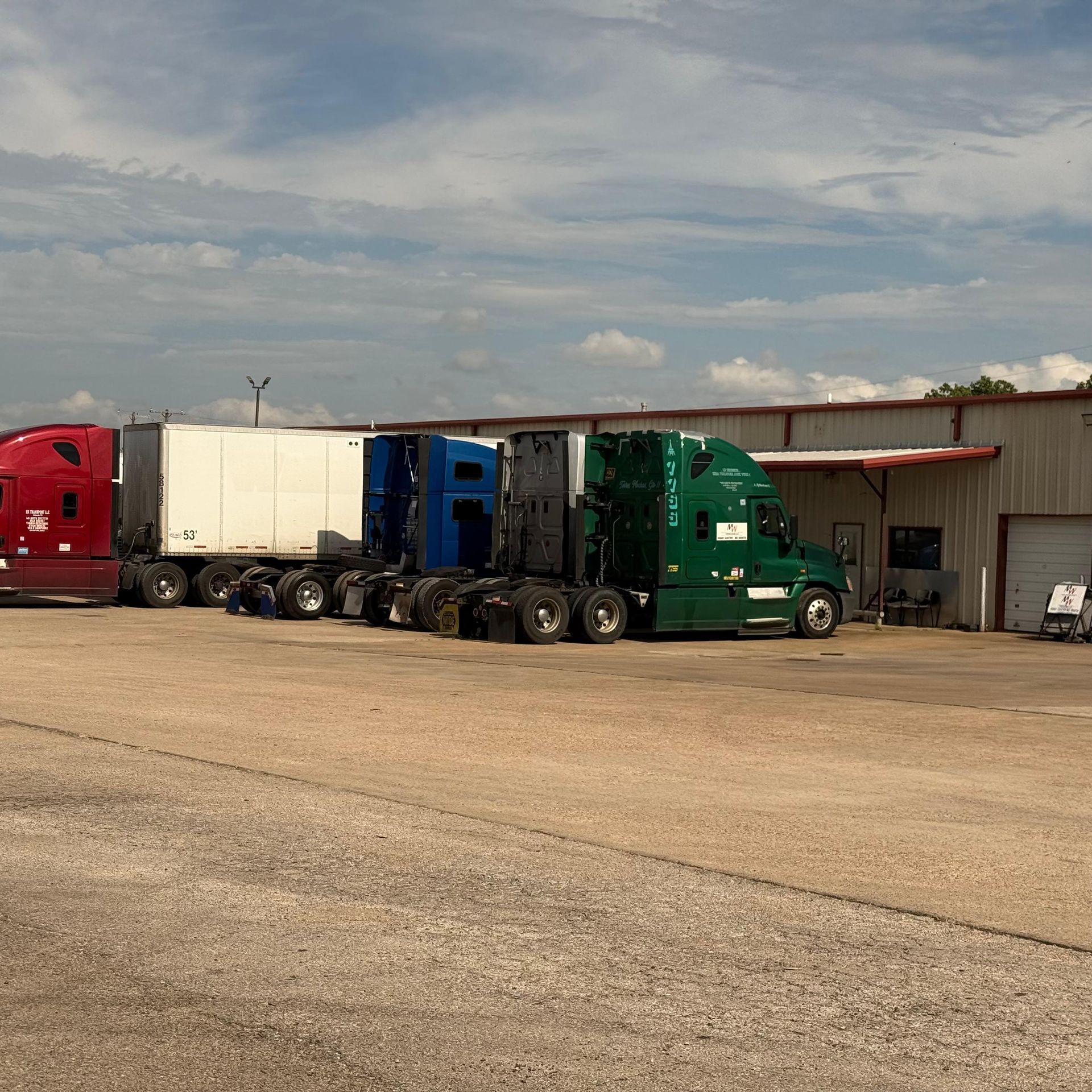 Semi-trucks of various colors parked outside a warehouse on a sunny day.