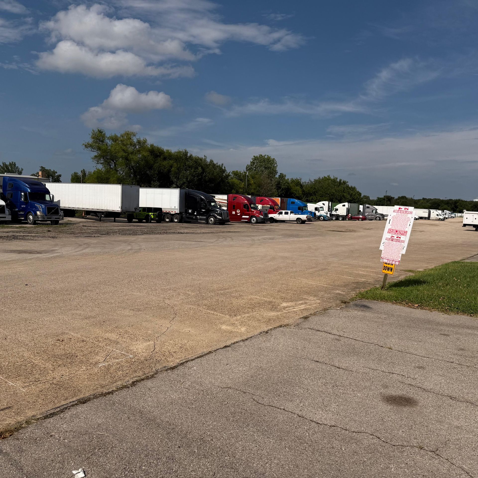 Semi-trucks parked in a gravel lot on a sunny day with a blue sky. A sign is in the foreground.