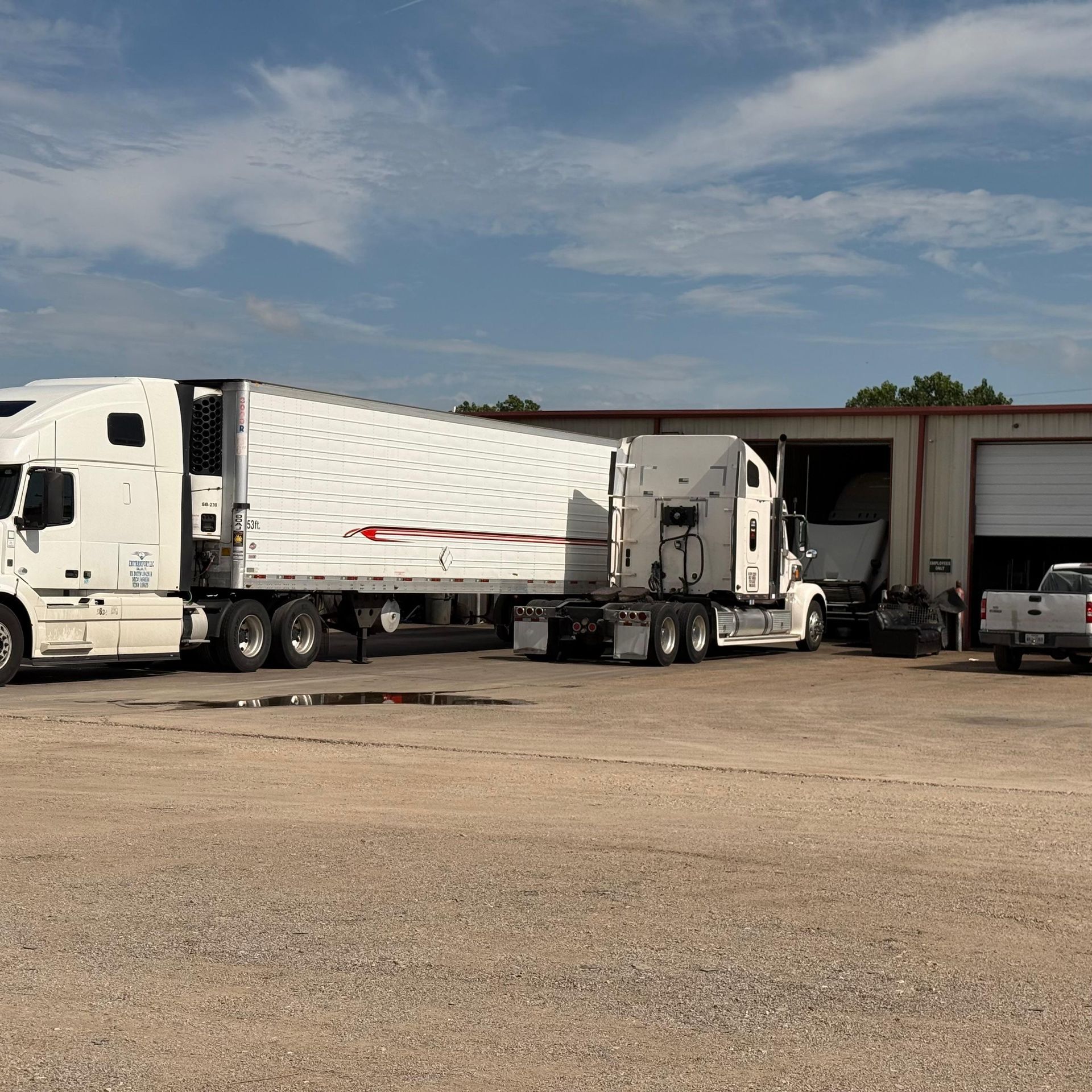 Two white semi-trucks parked near a building with an open garage door, under a blue sky.