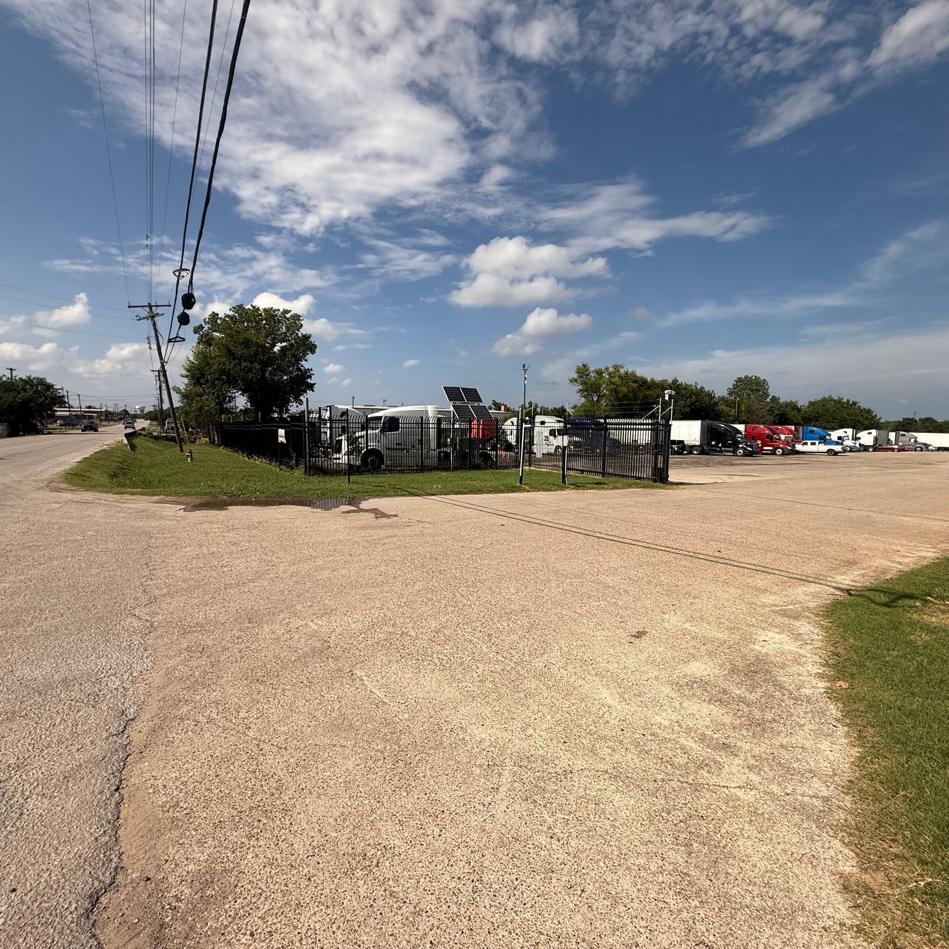 Gravel lot with parked trucks behind a fence, under a partly cloudy sky. Utility poles and a small tree are present.