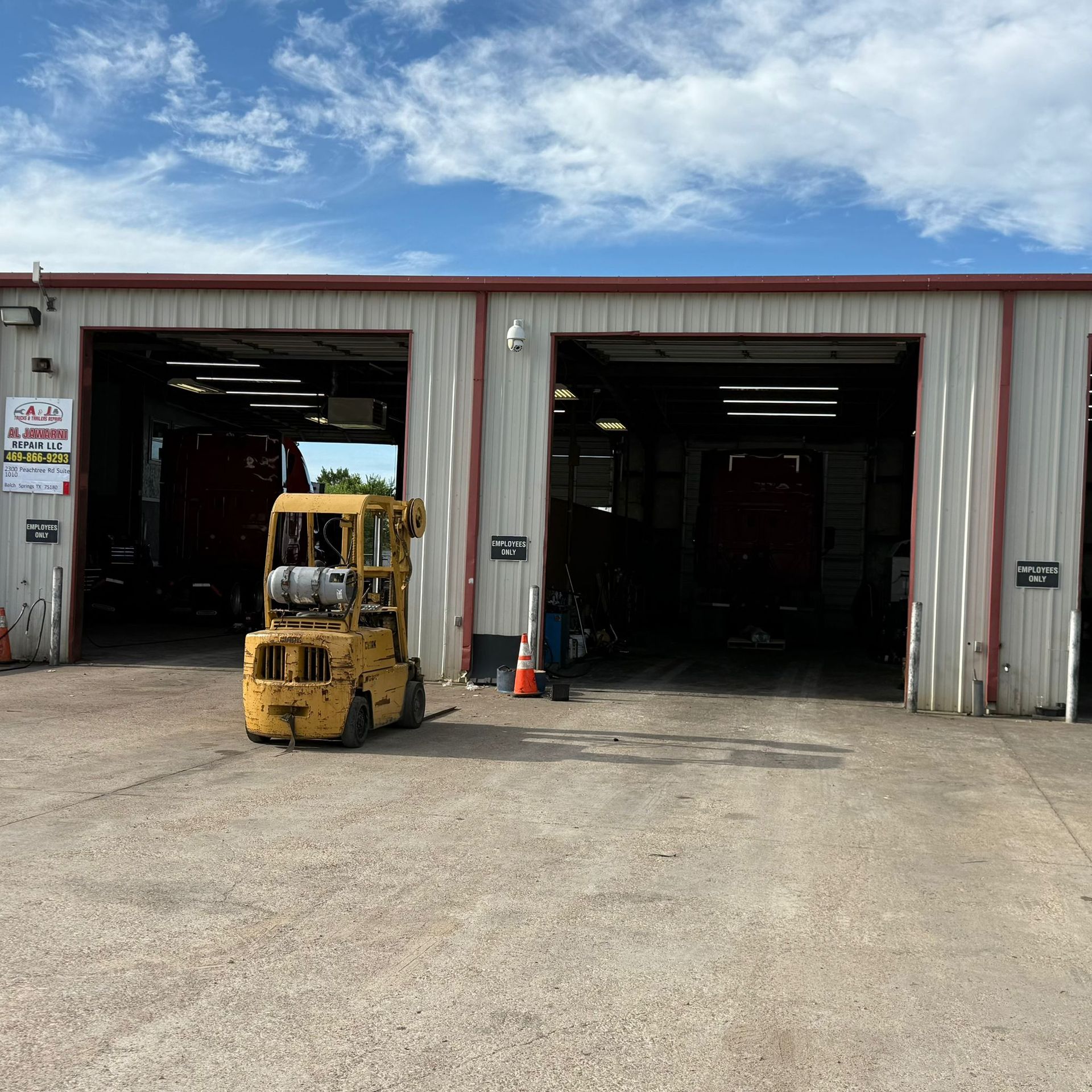 Yellow forklift parked in front of three open garage bays. Red trucks inside, cloudy sky.