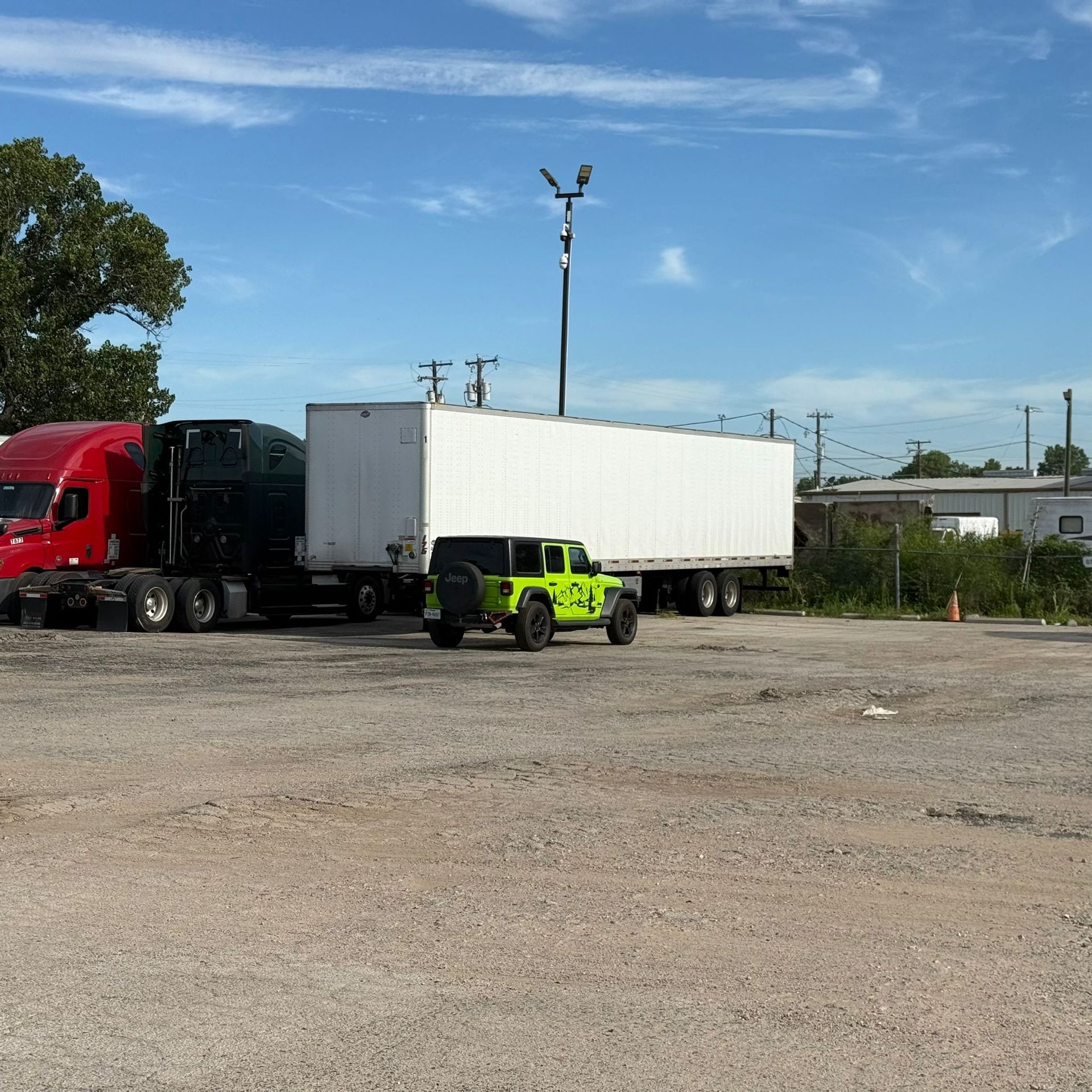 A green Jeep parked in a gravel lot next to semi-trucks and a white trailer under a blue sky.