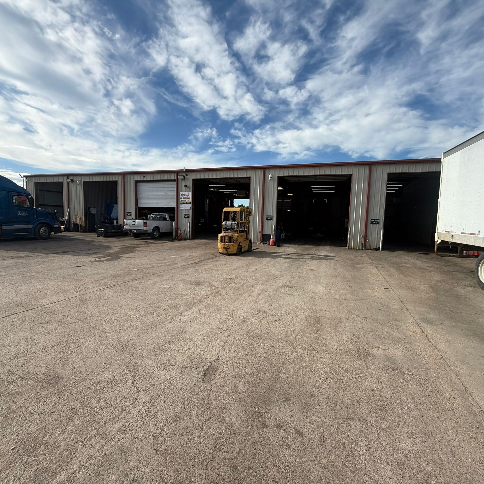 Exterior view of a truck repair shop with multiple bays, vehicles, and a forklift on a gravel lot under a cloudy sky.