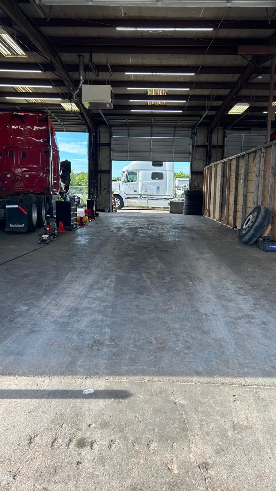 Inside a truck repair shop, looking out at a white RV. Red truck on the left, grey concrete floor.