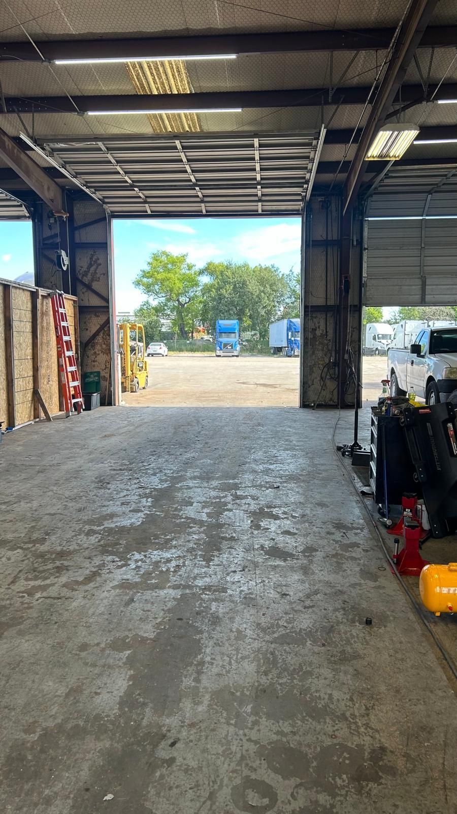 Open garage bay frames a view of a lot with trucks, trees, and a blue sky.