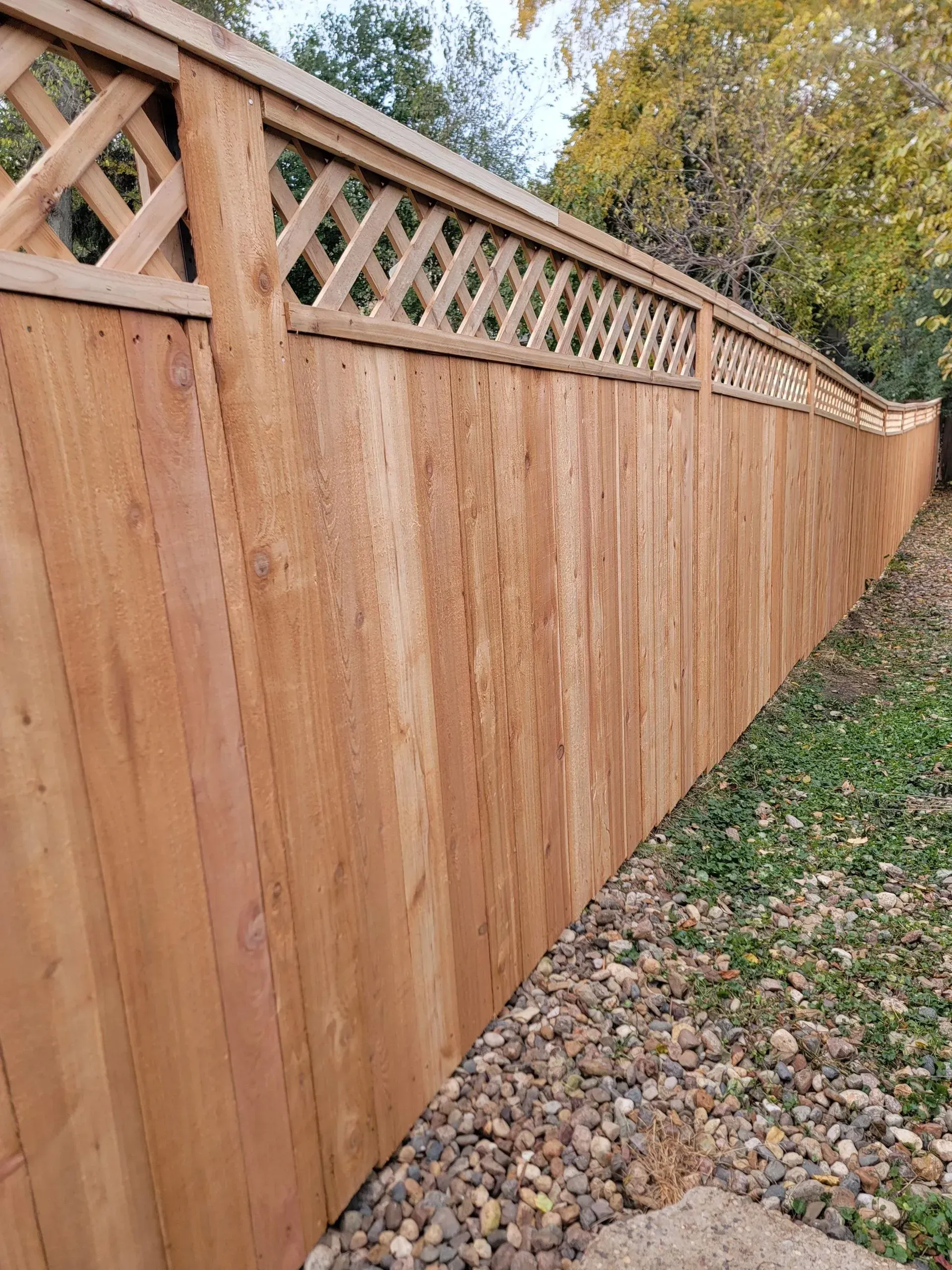 Wooden privacy fence with lattice detail, brown and tan, in a yard with gravel and grass.