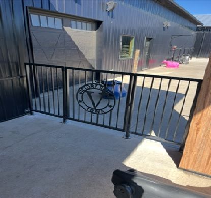 A black metal railing with a circular logo in the center stands on a concrete patio in front of a modern metal building.