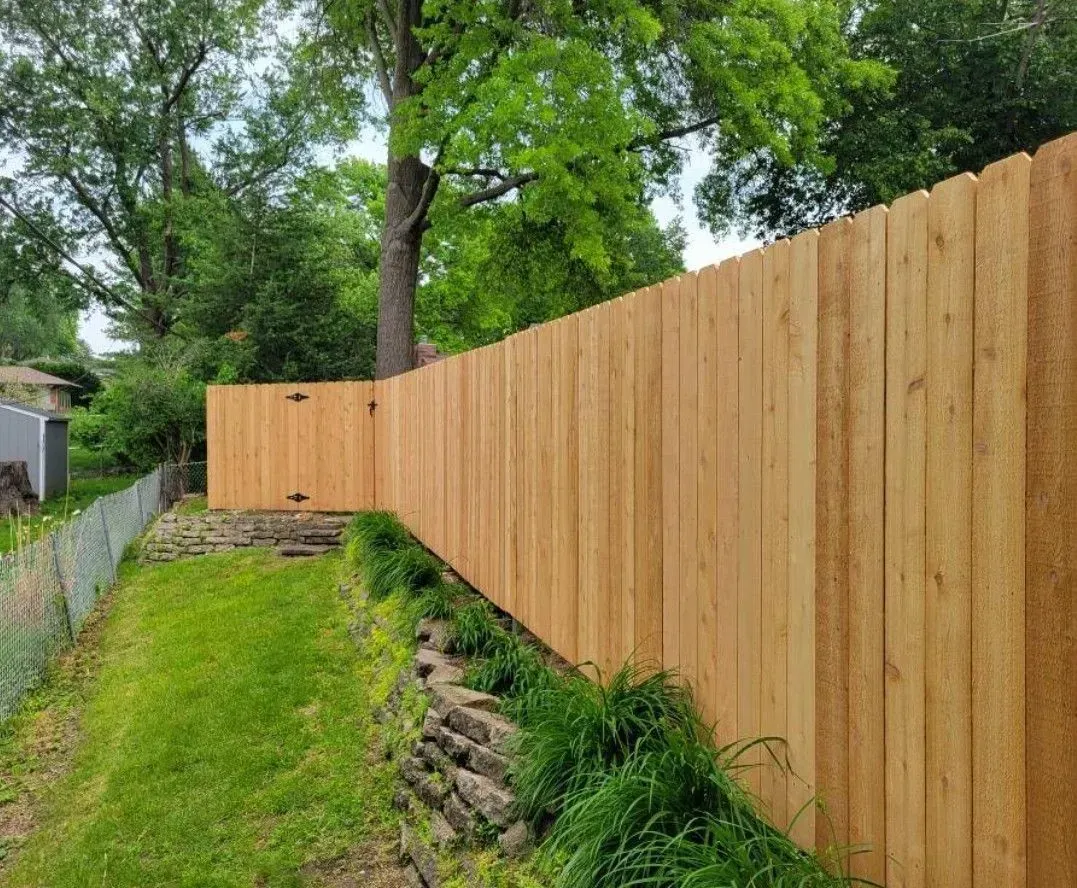 Wooden fence extending along a grassy yard, with a gate, trees, and a chain-link fence on the left.