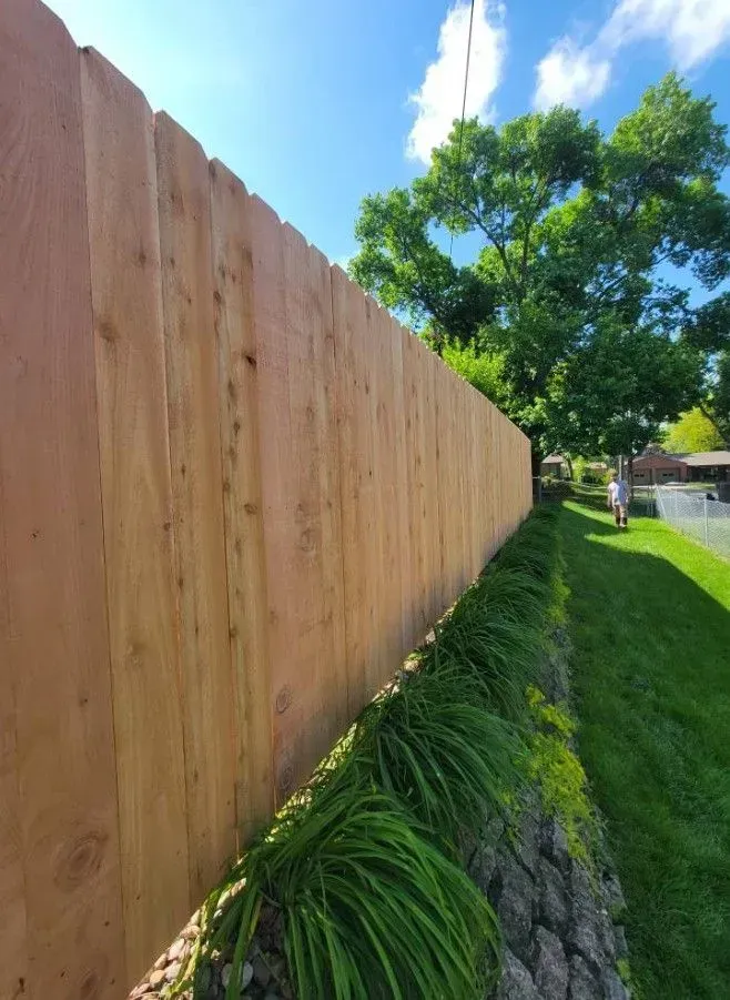 Wooden fence along a grassy slope, with green plants in front. A person is walking in the distance.
