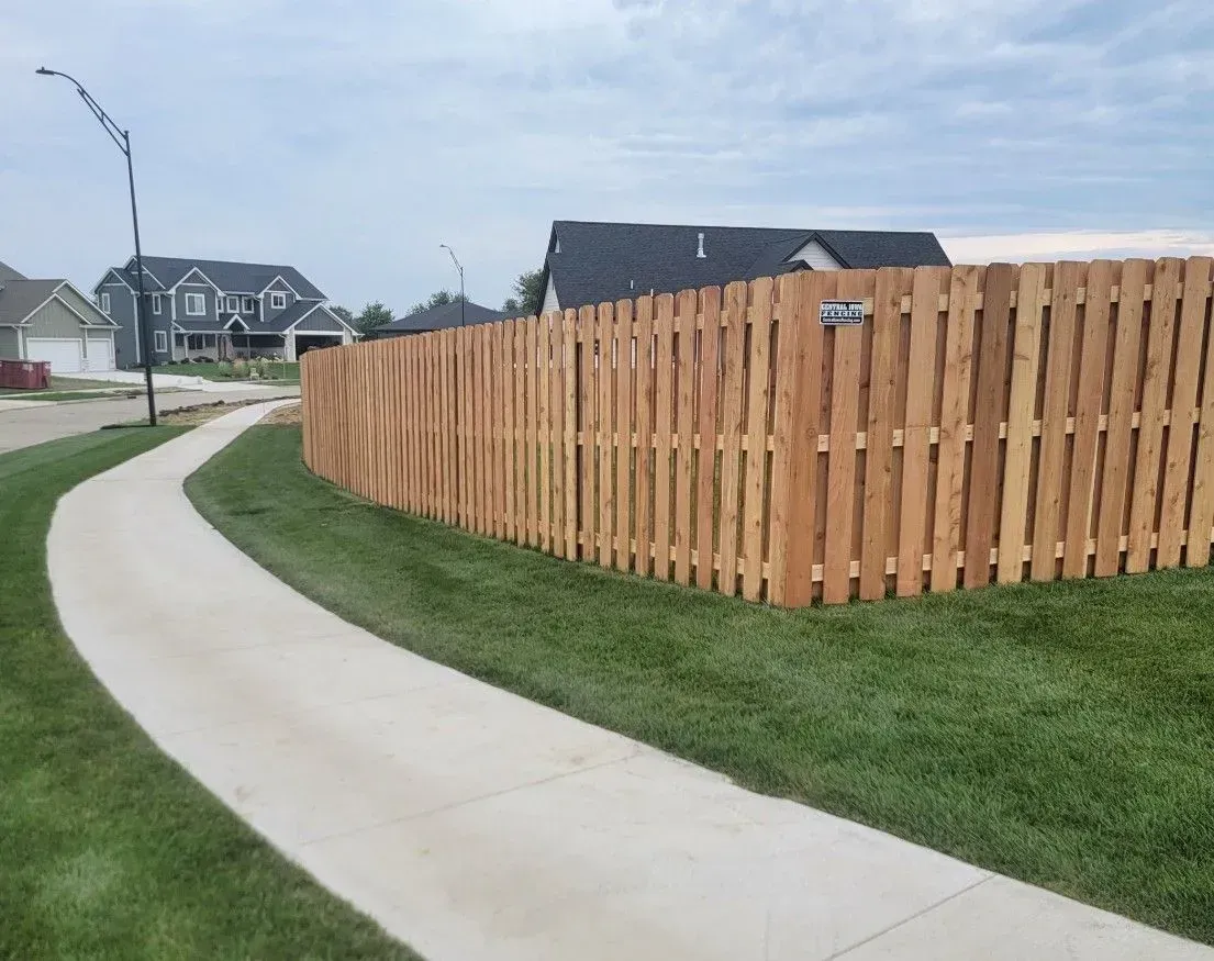 A wooden fence curves along a concrete pathway, bordered by green grass and houses in the background under a cloudy sky.