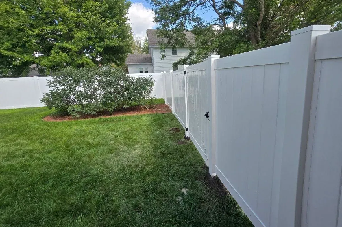 White vinyl fence encloses a green lawn, shrubs, and a house in the background on a sunny day.