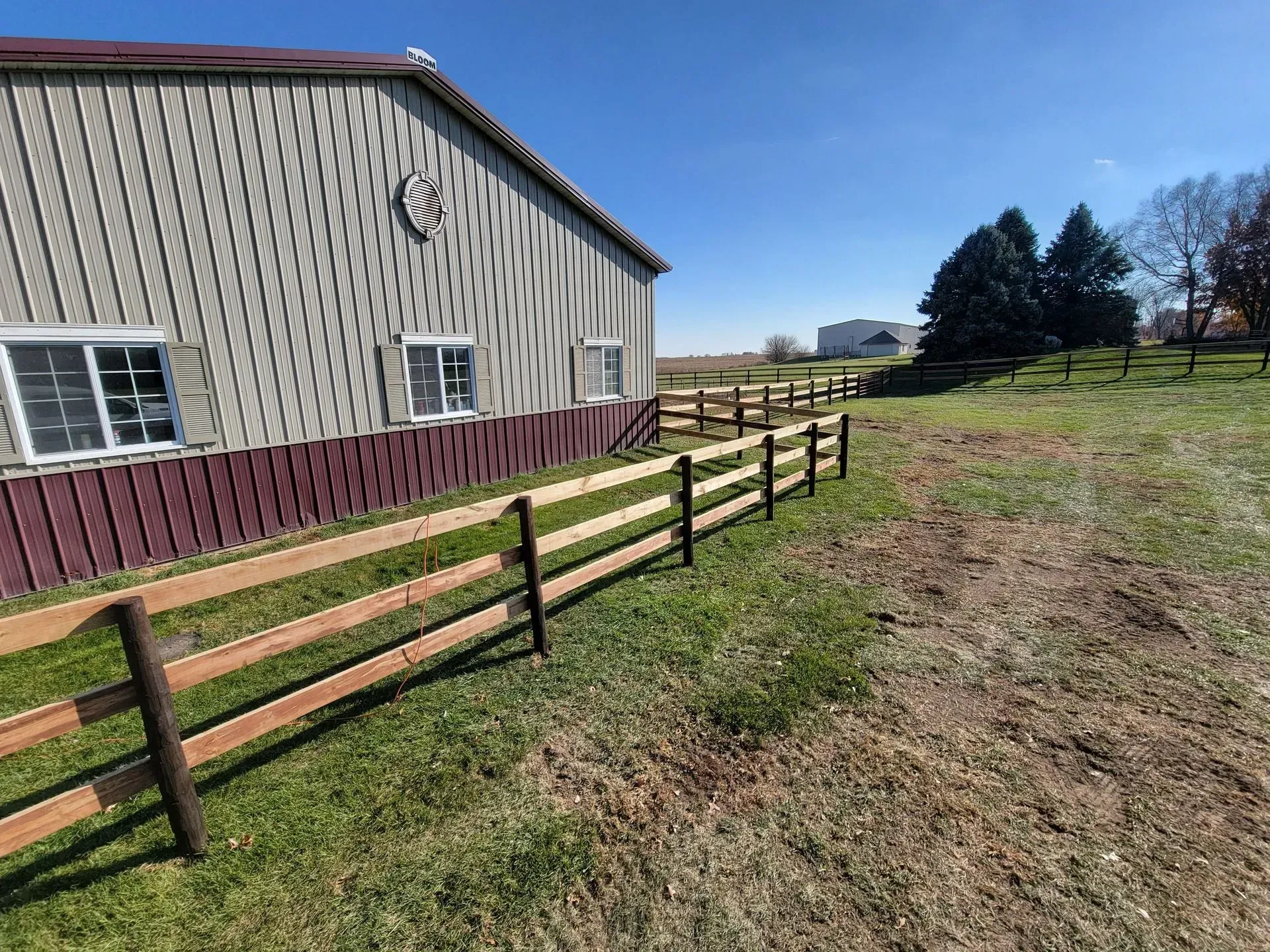 A wooden fence lines a field near a barn with a brown base and tan siding under a blue sky.