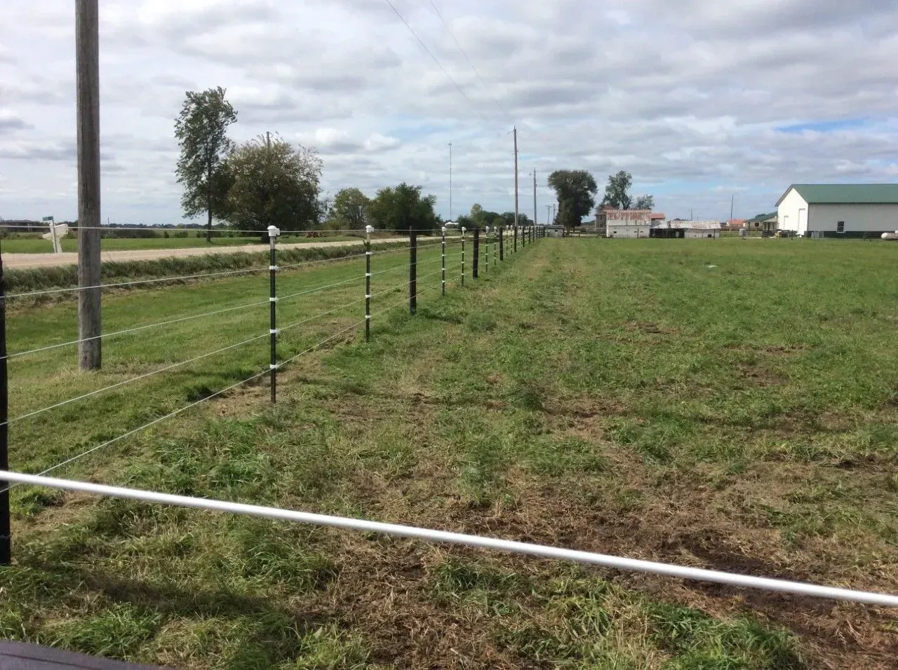 A grassy field with a fence. A white building and utility pole are in the background, under a cloudy sky.