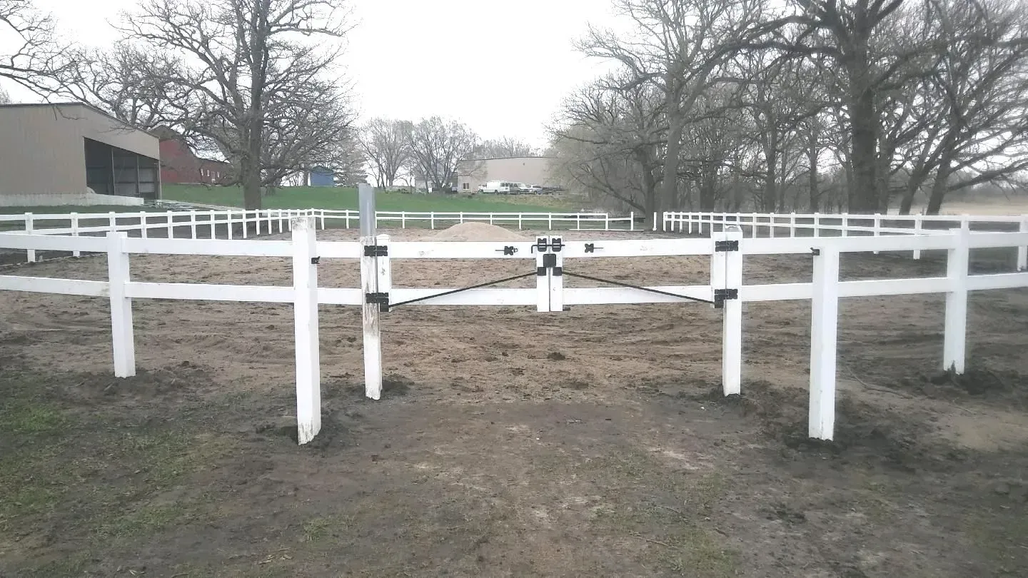White fenced-in riding arena with closed gate, dirt ground, and bare trees in the background.