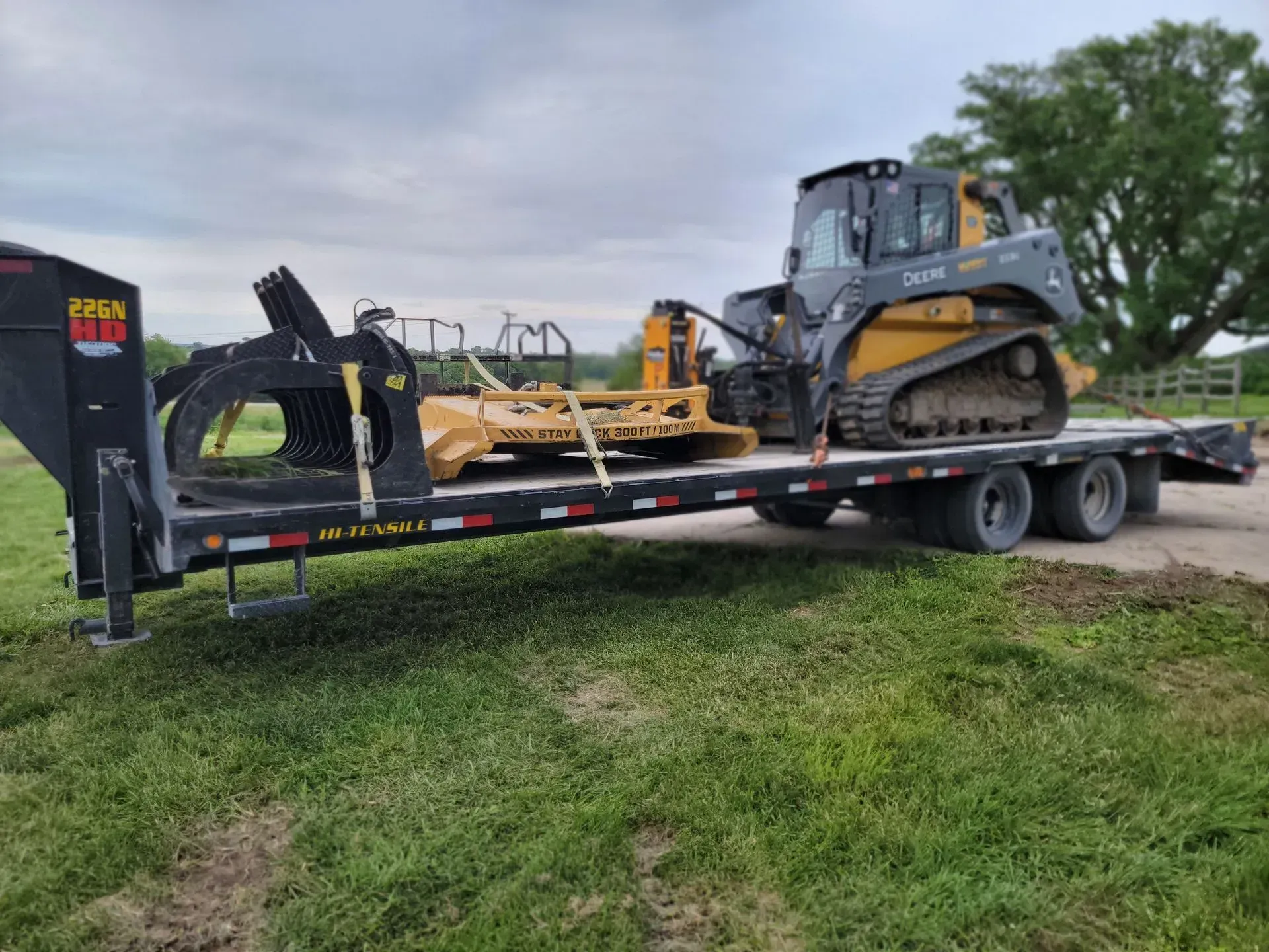 A John Deere skid steer with a brush cutter loaded on a black flatbed trailer in a grassy area.