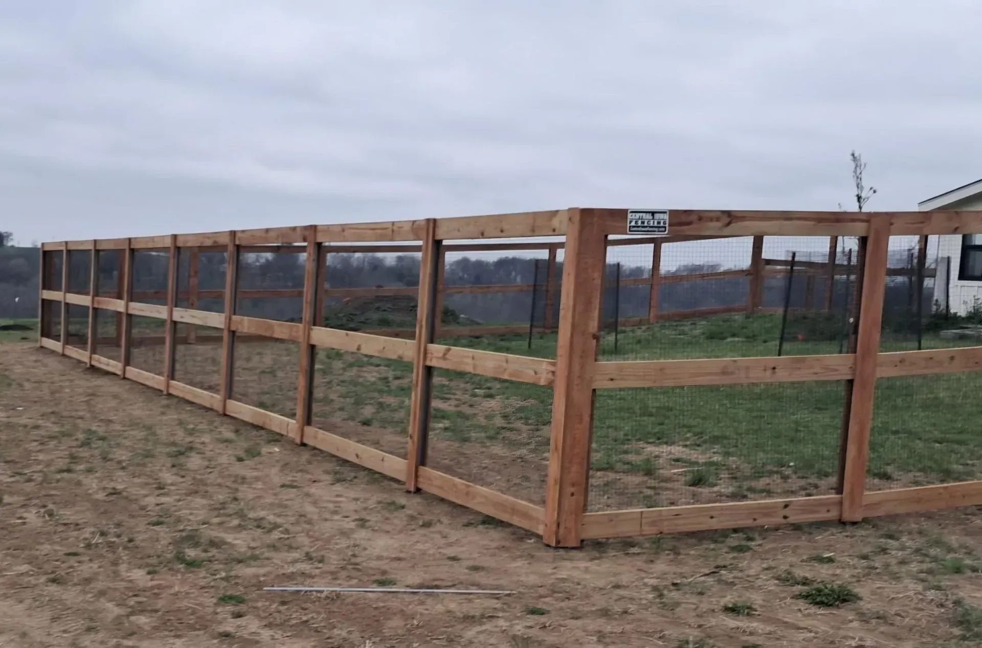 Wooden fence enclosing an area, with wire mesh, set in a grassy yard under an overcast sky.