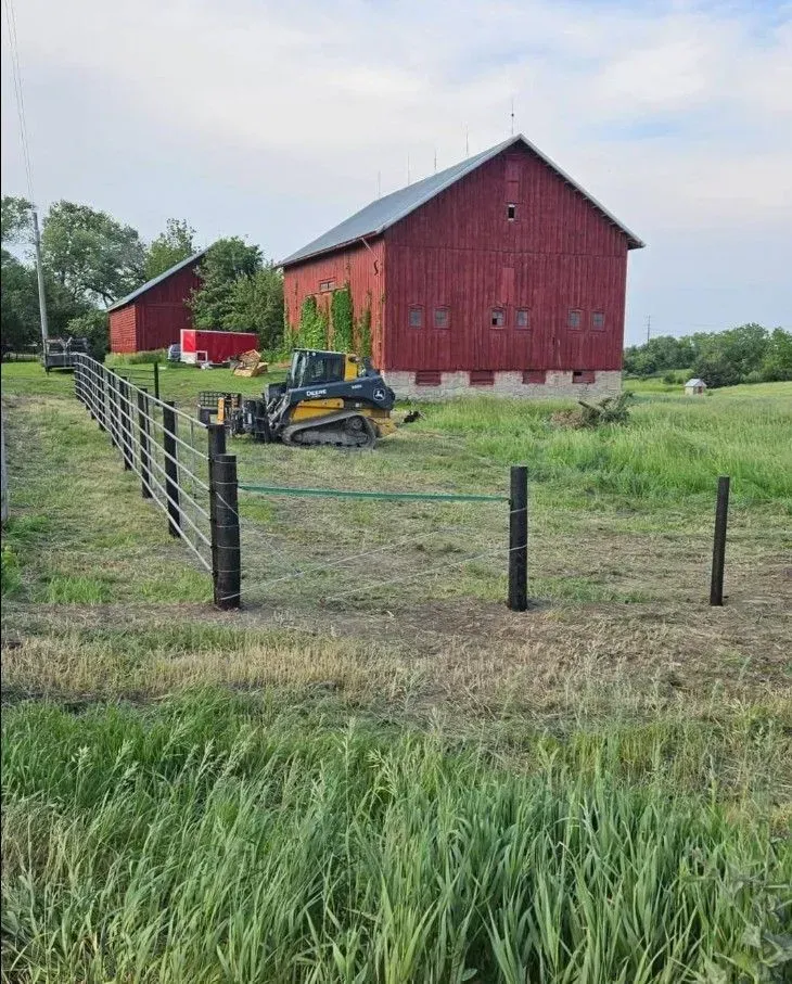 Red barns and fence construction in a grassy field; construction equipment nearby.