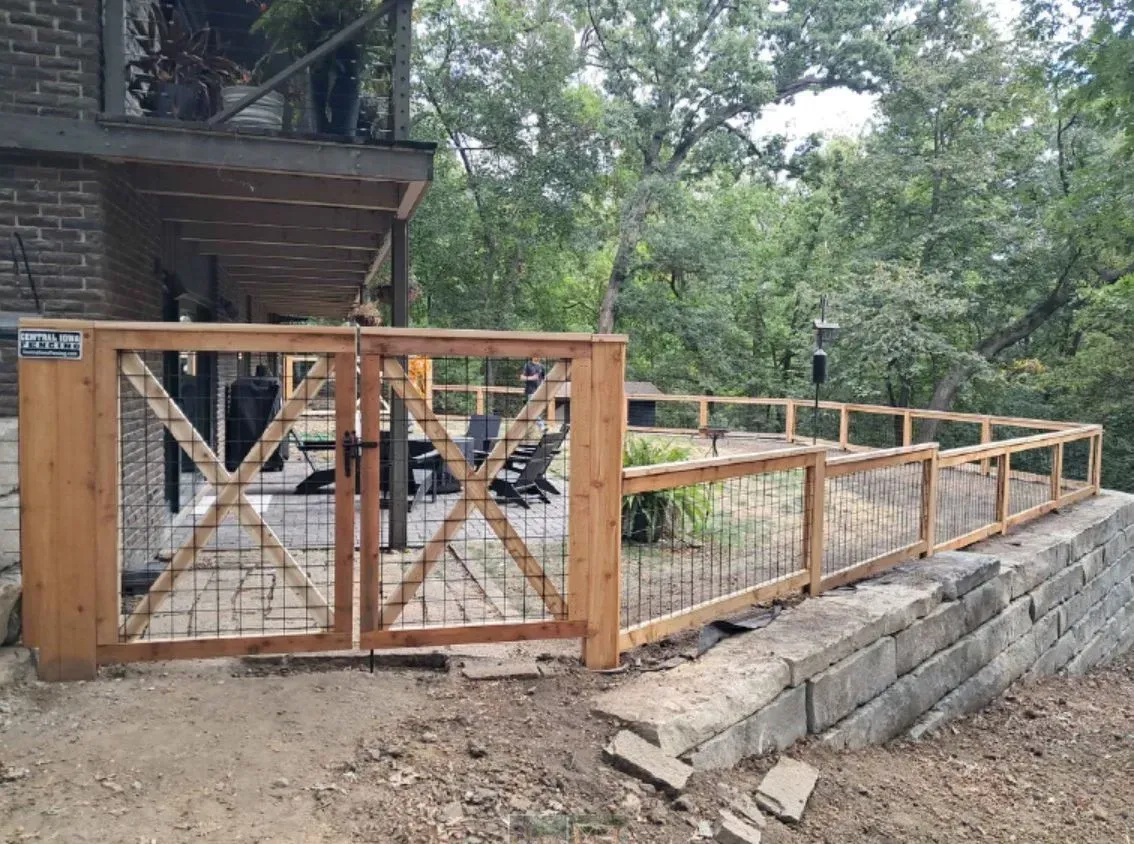Wooden fence with gate, bordering a deck and a stone retaining wall. Trees in the background.