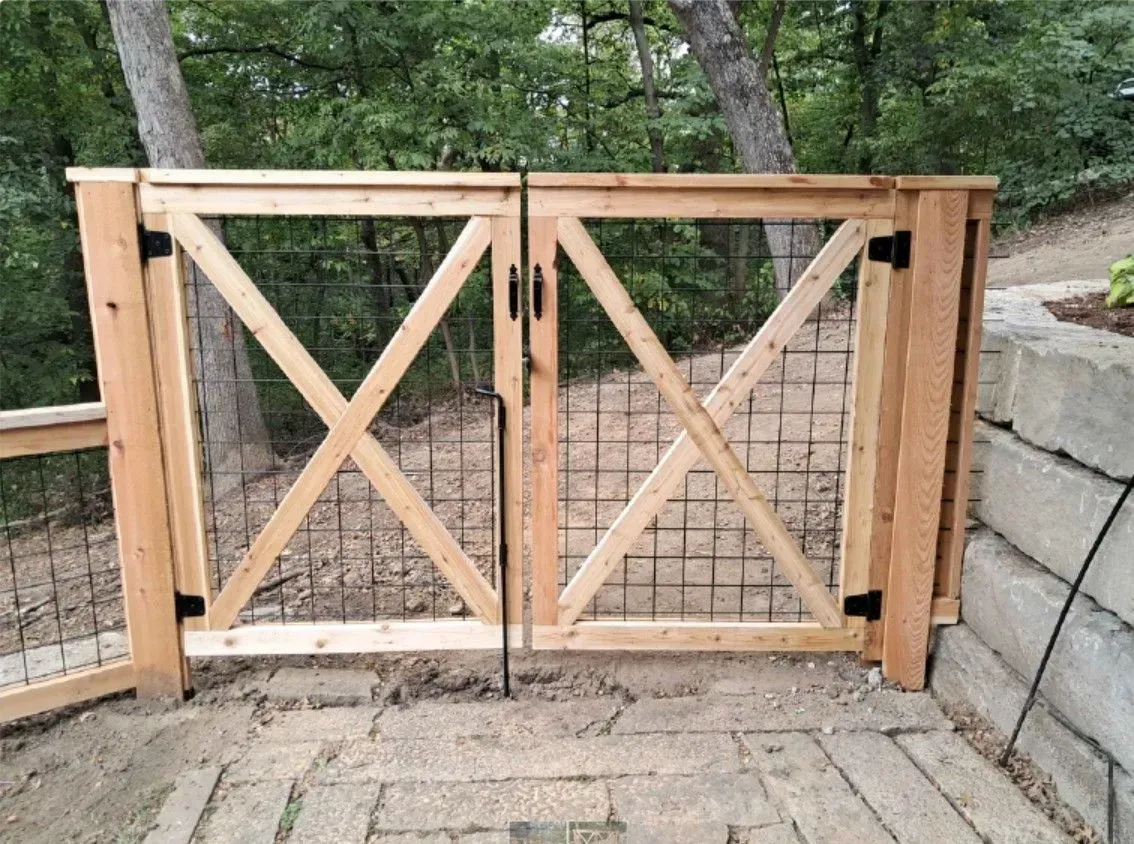 Wooden double gate with an X-shaped design, black hinges, and wire mesh on a paved patio.