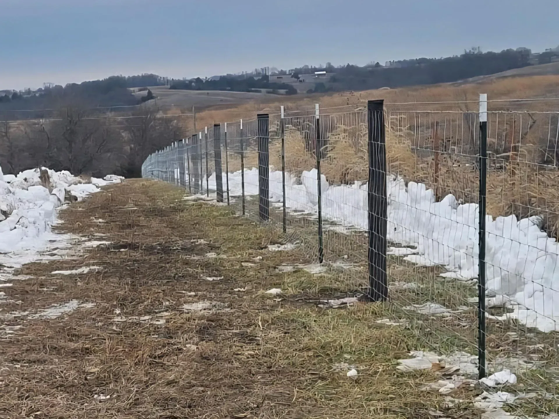 Fence line with snow along the edge in a rural field, with distant hills and a cloudy sky.