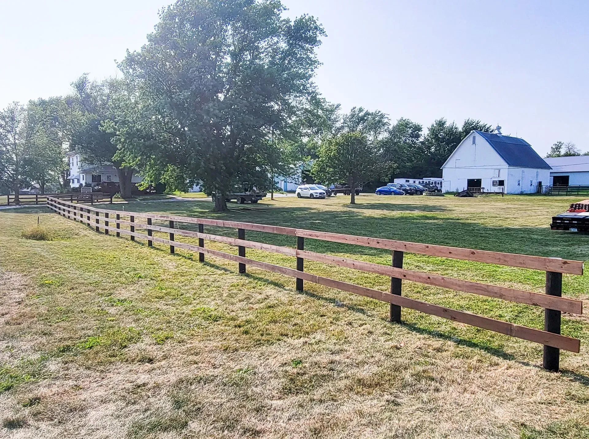 Two-rail wooden fence in a field, trees, white barn in background, sunny day.