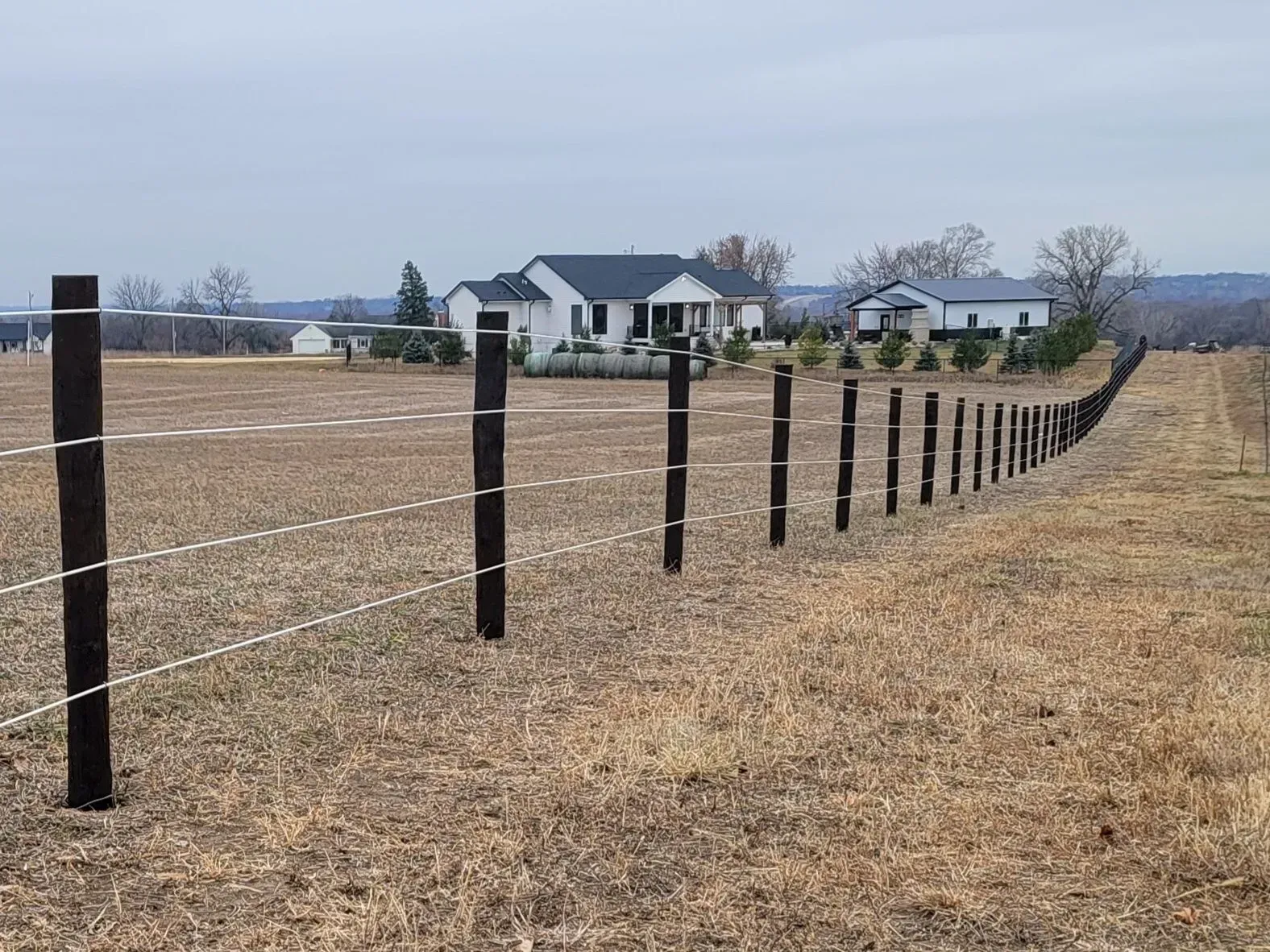Wooden fence with white electric wire in a field, houses in the background. Overcast sky.