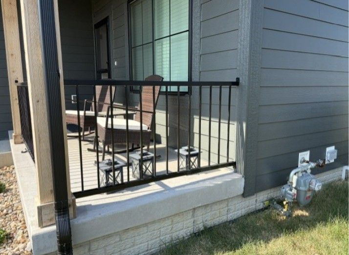 Small porch with metal railing, chairs, and a grill beside gray house siding