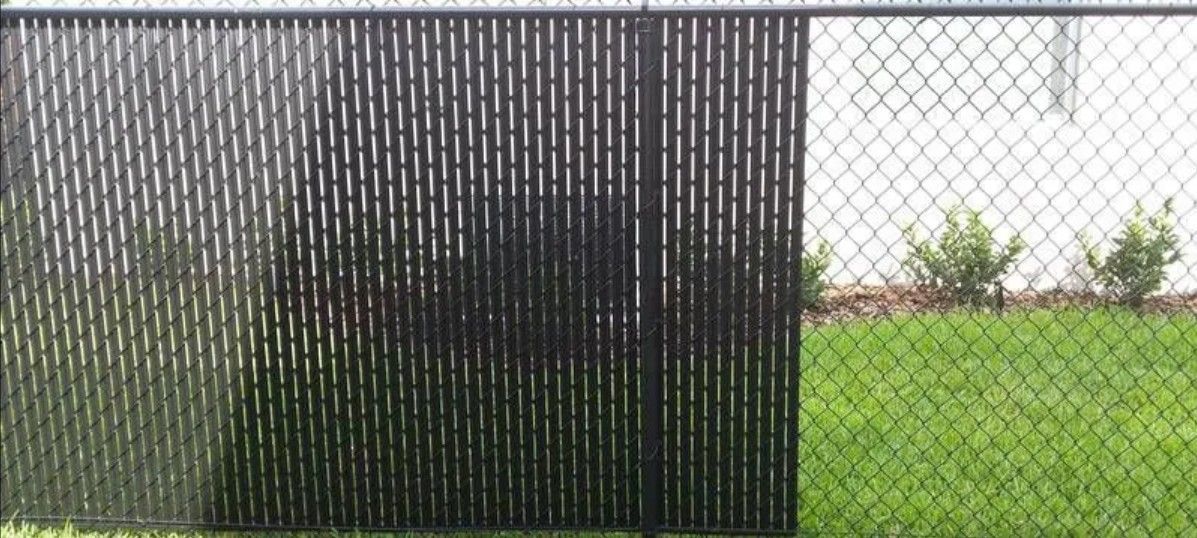Chain-link fence with black privacy slats on the left side, with green grass and shrubs in front of a white wall.