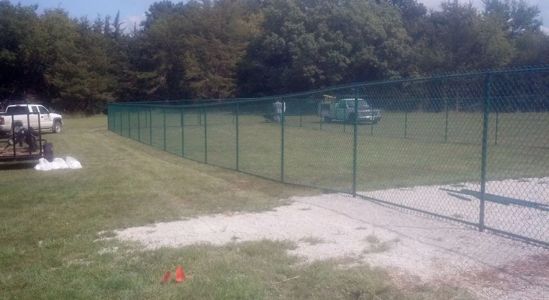 Green chain link fence bordering a gravel driveway and grassy area, trees in the background, parked vehicles visible.