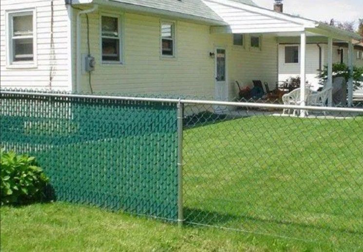 Green chain-link fence with green privacy slats in a backyard, beside a light-yellow house and lawn.