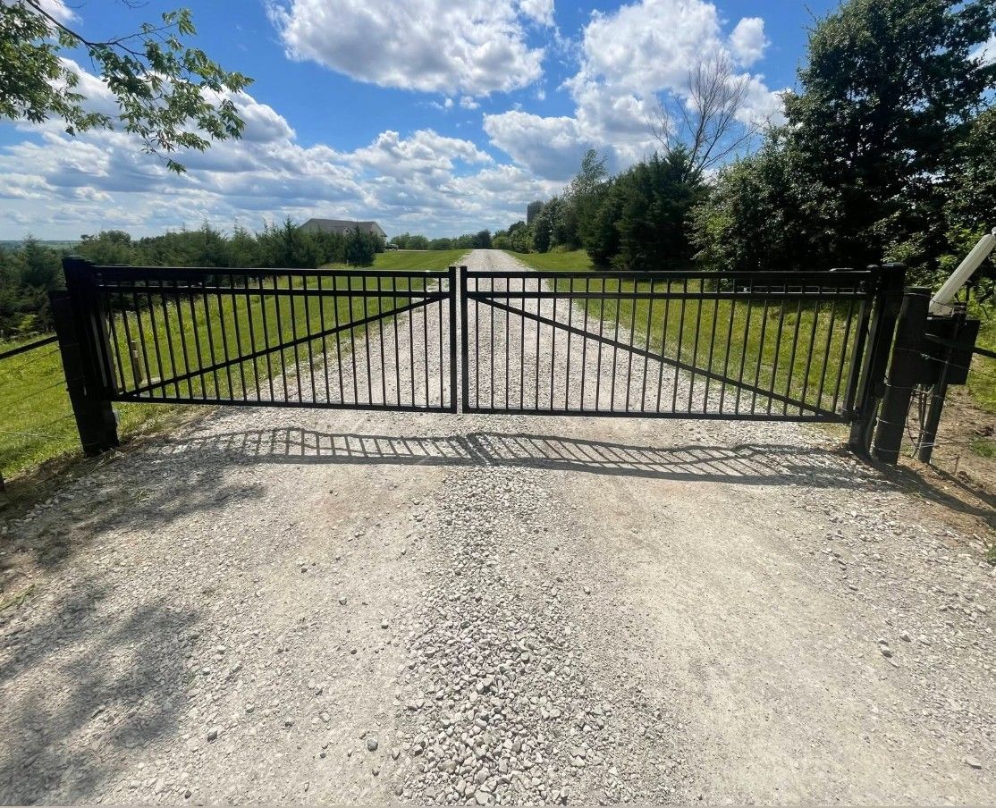 Black metal automatic gate across a gravel driveway under a blue, cloudy sky, with trees and grass on both sides