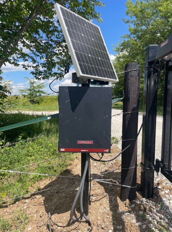 Solar panel mounted on a black utility box outdoors beside an automatic gate and trees.
