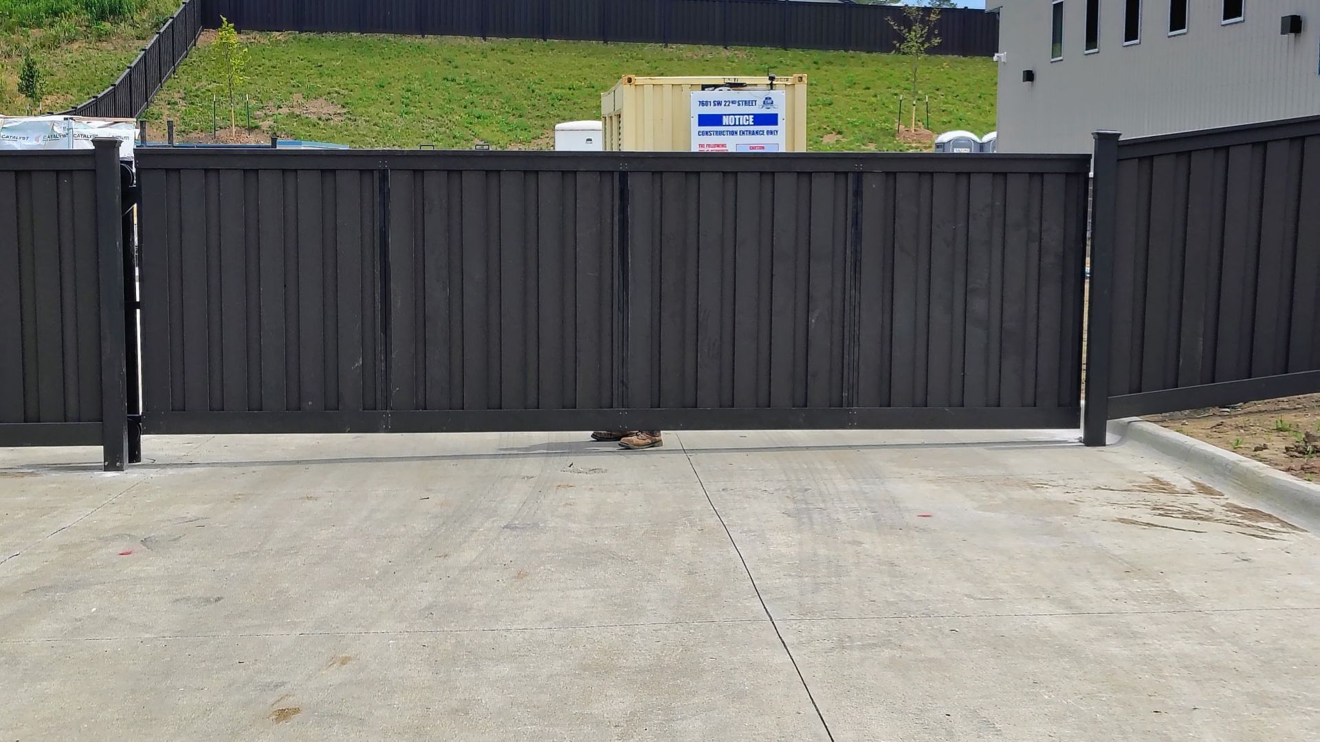 Black metal sliding automatic gate blocking a concrete driveway, with a grassy slope and buildings behind it.