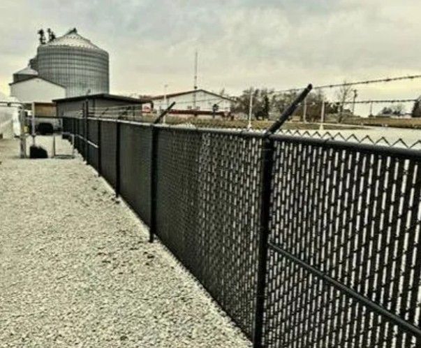 Black chain-link fence topped with barbed wire, bordering a gravel path and industrial buildings.