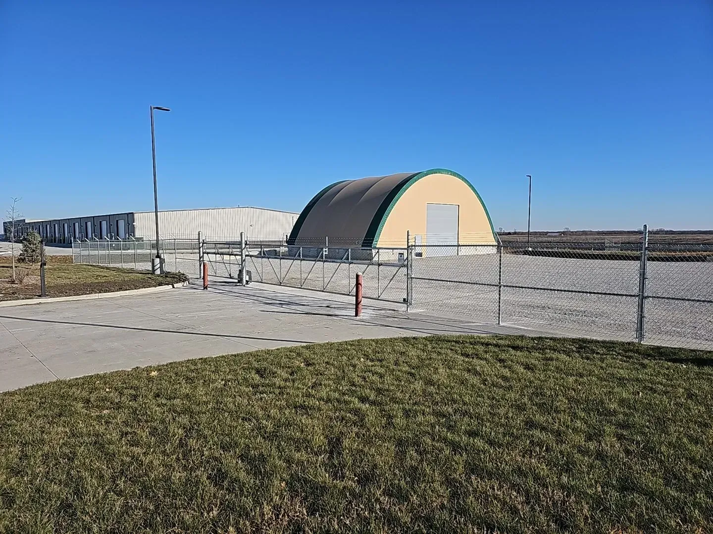 Quonset hut with beige walls, green trim, and a closed door, behind a chain-link fence on a sunny day.