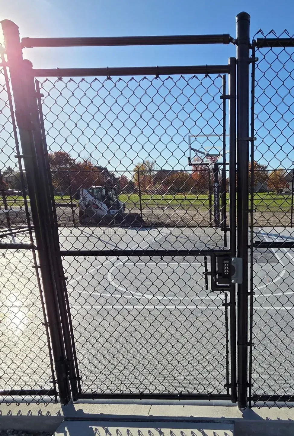 Black chain-link fence with gate, enclosing a concrete basketball court. A small tractor is visible in the background.