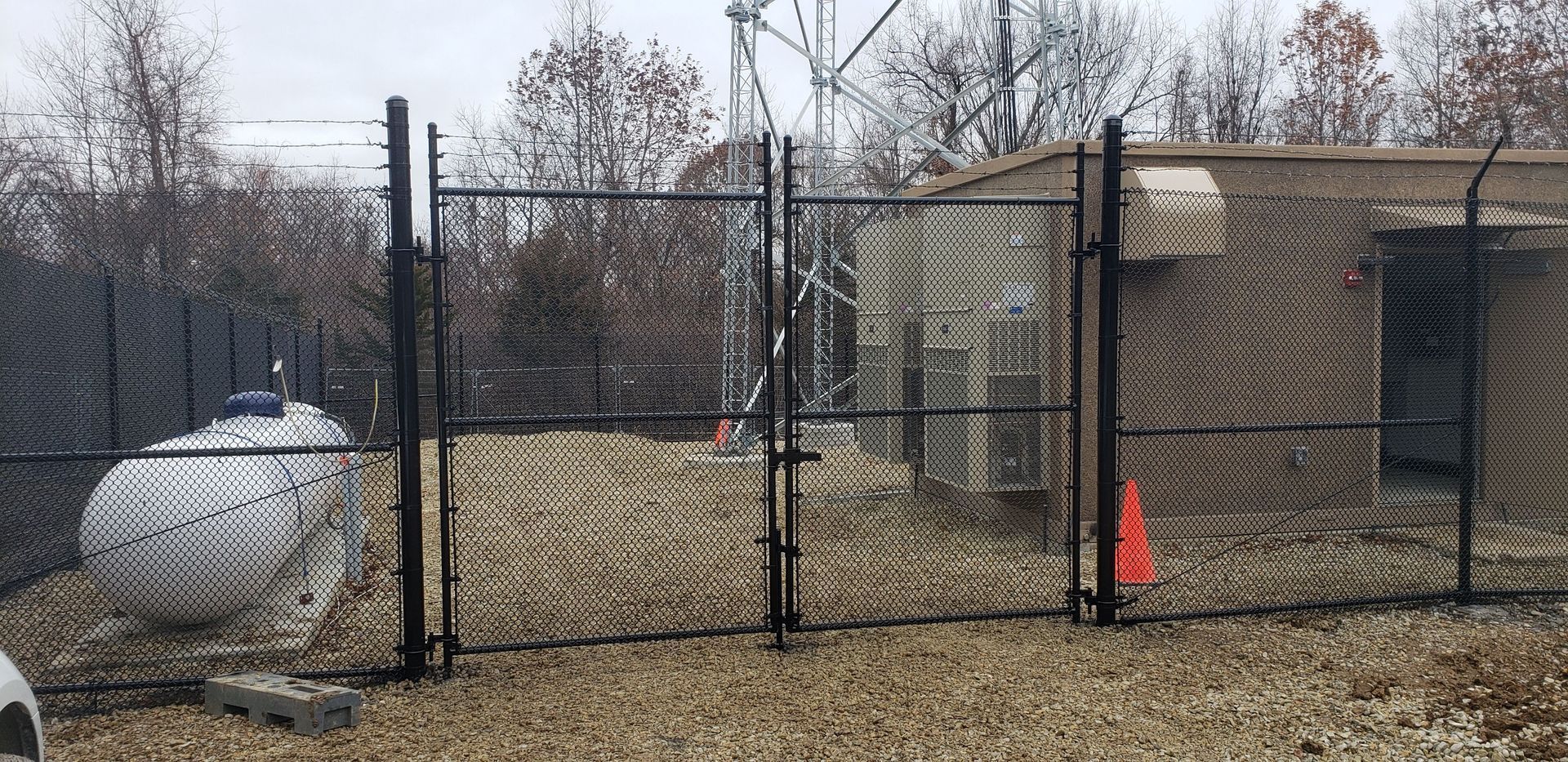 A fenced industrial area, showing a gate, a tank, and a building with an antenna tower visible in the background.