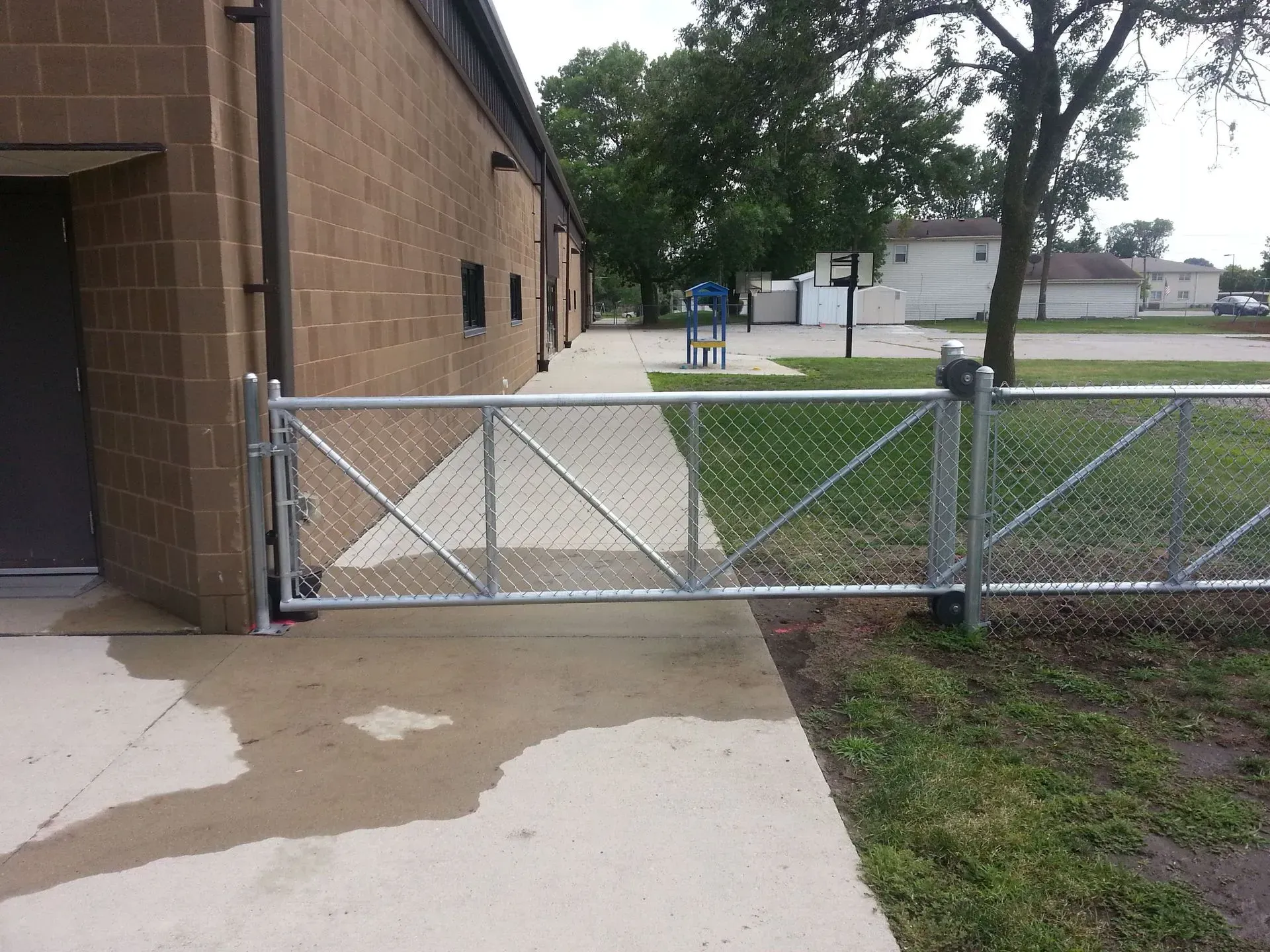 Chain-link fence with gate blocks a concrete walkway next to a brick building and grass.